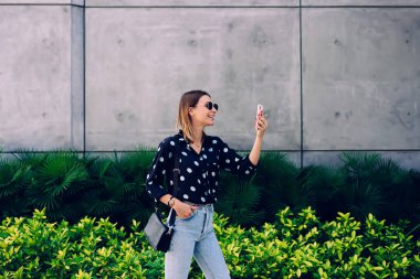 Sıradan kıyafetler ve güneş gözlükleriyle gülümseyen şık genç bir bayanın şehir caddesinde el ele dururken akıllı telefonuyla selfie çekmesi yan görünüm.