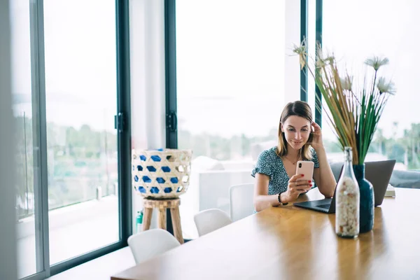 Smiling prosperous blonde woman in home interior reading messages on mobile phone during distance job, happy caucasian female blogger satisfied with online income and project working remotely