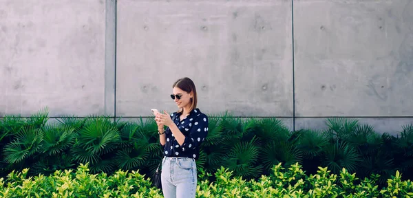 夏の晴れた日に街の通りに立っている間 スマートフォンを使用してカジュアルな服やサングラスで幸せなスタイリッシュな若い女性 — ストック写真