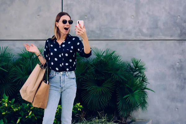 ショッピングバッグで植物の壁の近くに立っているスタイリッシュなカジュアルな服の若い女性を驚かせ 口を開けてジェスチャーで手を動かす — ストック写真