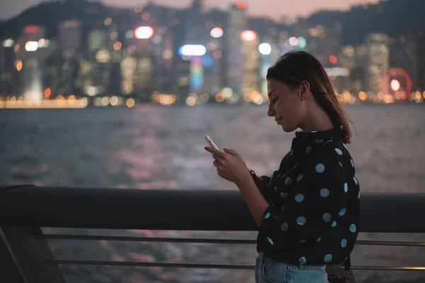 スマートフォンでネットサーフィンをしている夜の街の明かりを背景に静かに立つ流行の女性の笑顔の側面図 — ストック写真