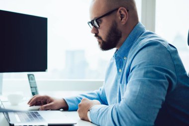 Resmi tişörtlü ve gözlüklü, modern ofiste oturan ve kripto para ticareti yapan bitcoin planıyla çalışan kendine güvenen erkek çalışanın yan görüntüsü.