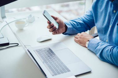 Yukarıdan gelen resmi olarak tanınmayan erkek, modern ofisteki dizüstü bilgisayarda çalışırken akıllı telefondan sohbet ediyor.