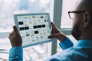 Pencere kenarında otururken mavi gömlek ve gözlük takan, dijital tablet kullanan tanınmayan erkek sakallı çalışanın arka plan görüntüsü