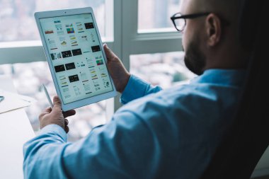 İş yerinde pencerenin yanında otururken tablet ve dijital kalem kullanan resmi kıyafetler ve gözlük takan sakallı adamın arka planı