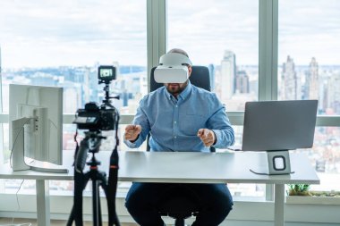 VR gözlük takan erkek blogcu ofis alanındaki dijital kameraya video kaydederken pencerenin yanındaki bilgisayarlarla masada oturuyor