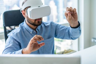 Modern ofiste resmi tişört giyen erkek ofis çalışanı kollarını kaldırmış masada otururken VR gözlükleriyle oyun oynuyor.