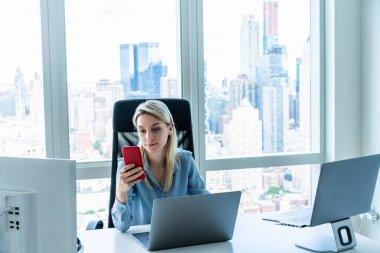 Modern akıllı telefon kullanan, uzun sarı saçlı sakin bayan çalışanı masadaki modern ofiste bilgisayar ve pencerenin yanındaki dizüstü bilgisayarlarla çalışırken...