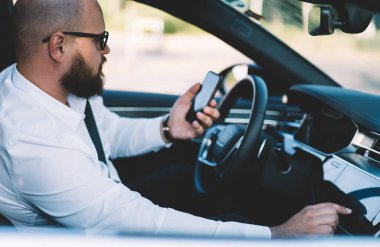 Lüks bir araçta oturan ve araba teybine bakarken cep telefonunda sörf yapan şık giyimli serbest çalışan erkek.