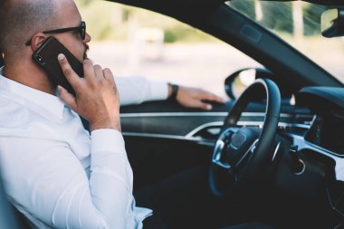 Gözlüklü bir erkek girişimcinin güneşli bir günde açık pencereli bir arabada otururken telefonla konuşmasının yan görüntüsü