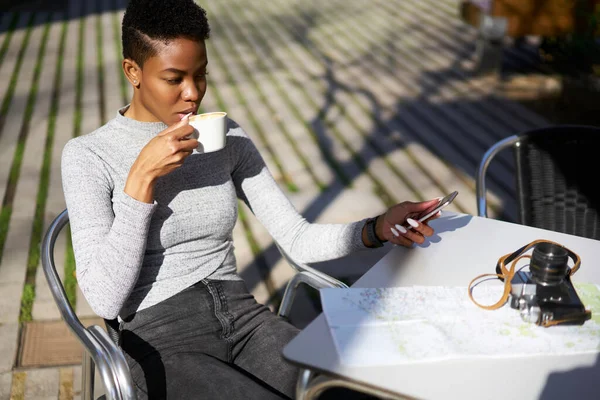 作物の若い深刻なアフリカ系アメリカ人の女性旅行者の上から写真カメラと紙の地図で屋外のカフェのテーブルに座っている間 スマートフォンや飲酒コーヒーの画面を見てカジュアルな服装で — ストック写真