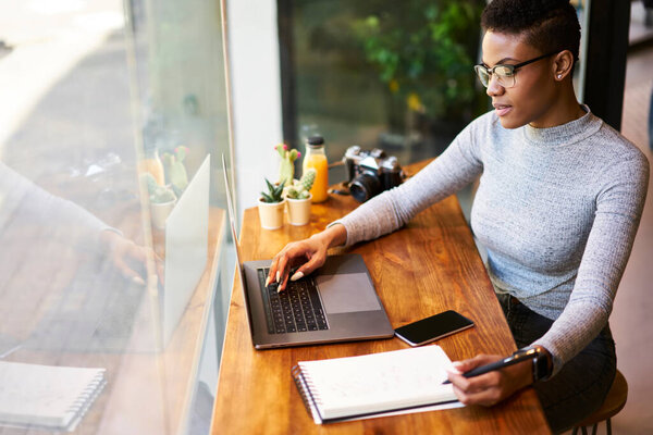 From above of serious African American female journalist sitting in cozy workspace while working online on new article for magazine