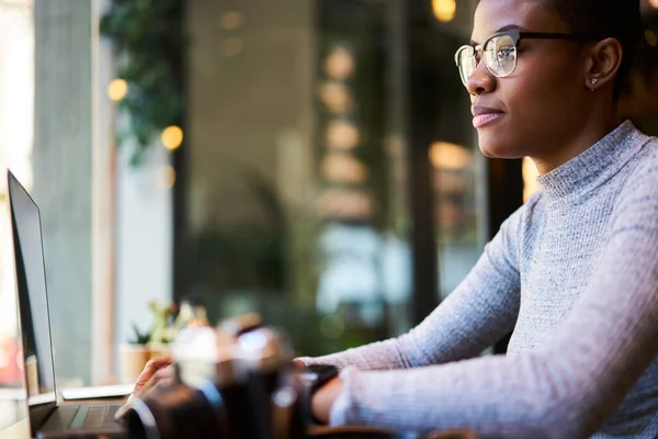 身穿休闲装和眼镜的年轻非洲裔美国博主与上网本坐在桌旁 在咖啡店工作的侧视图 — 图库照片