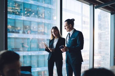 Modern işyerindeki büyük pencerenin önünde duran resmi kıyafetli genç iş adamları yeni projeyi değerlendirirken tablete dikkat ediyorlar.
