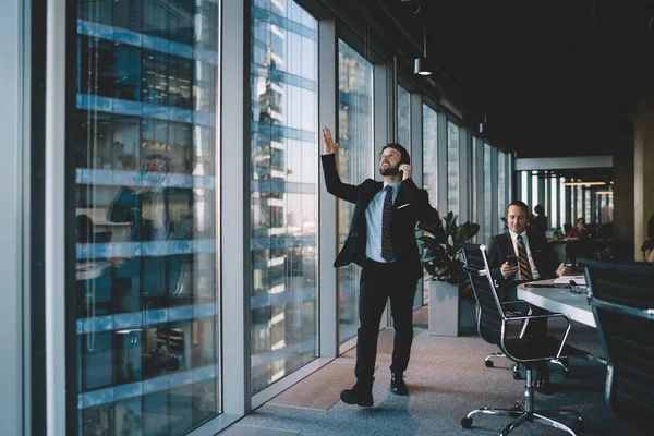 Annoyed businessman in classy wear walking along modern office and speaking on mobile phone while expressing anger and gesticulating on background of busy colleagues