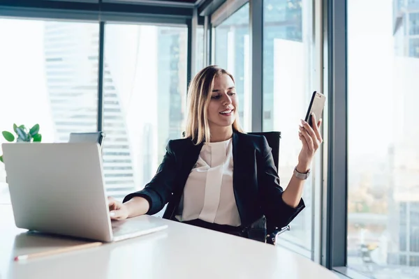 穿着雅致服装的女企业家坐在办公室桌子旁 一边浏览手机 一边在上网本上打字 一边为新项目工作 — 图库照片