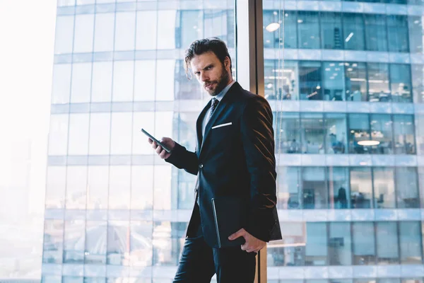 携帯電話でメッセージを送信し カメラを見ている間 現代の職場で窓の近くにノートブックを持つ上品なスーツ姿の男性起業家の眉の下から — ストック写真