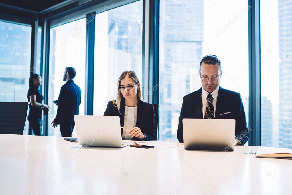 Concentrated man and woman in formal elegant suits sitting at white conference desk and typing on netbooks working in big open space office