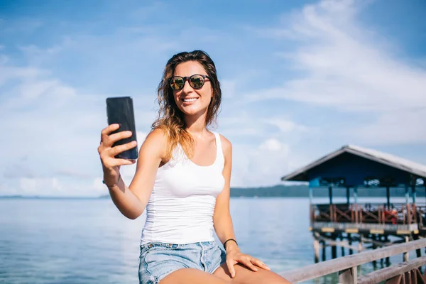Cheerful woman in sunglasses writing email message sharing impressions about solo travelling on Maldive, happy female tourist using roaming connecting for social networking via smartphone application