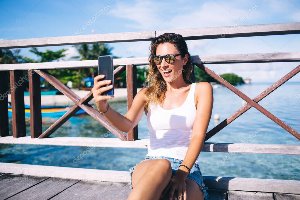 Prosperous female tourist in sunglasses shooting positive live streams for tell about Philippines vacations, cheerful girl using modern smartphone device for making selfie content from Hawaii