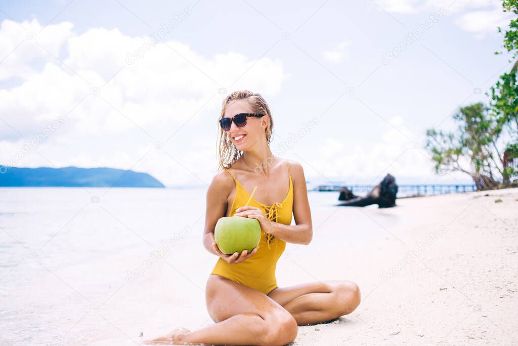 Happy Caucasian woman with exotic fruit sitting at white sand of Maldives and smiling, pretty female swimmer holding coconut beverage enjoying free time for sunbathing on Bahamas coastline