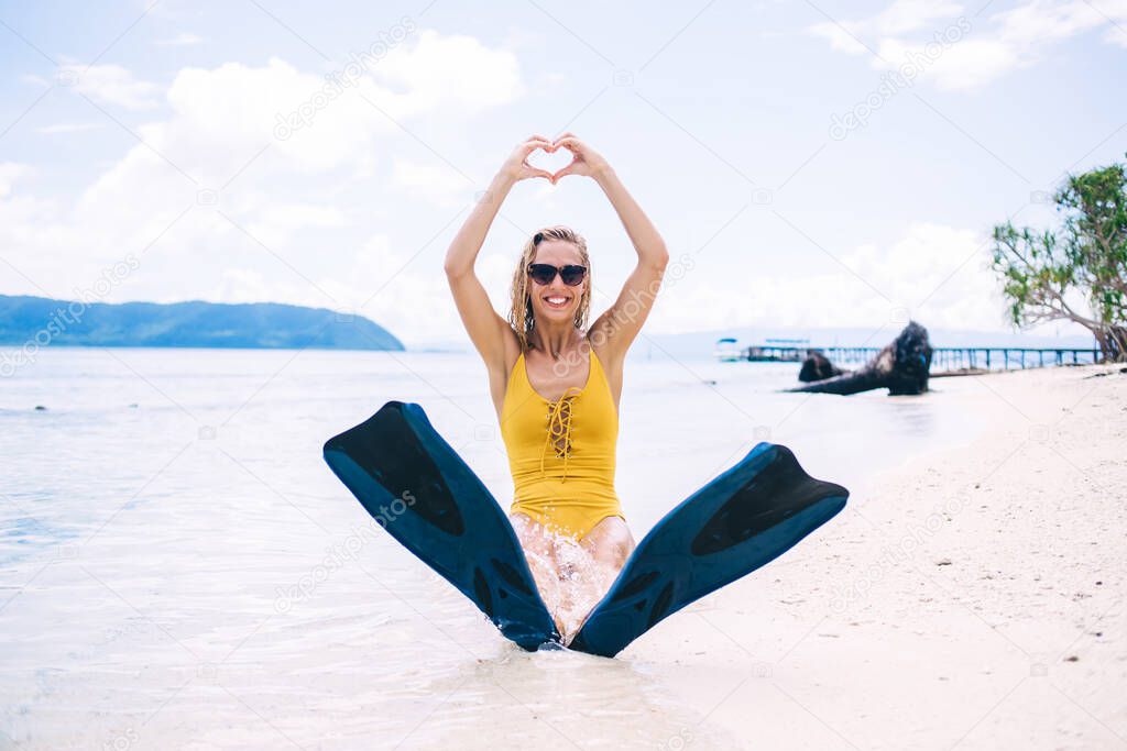 Portrait of funny female swimmer in flippers sitting at white sand beach and showing heart over head, cheerful Caucasian woman in sunglasses enjoying summer vacations for visiting tropical Hawaii