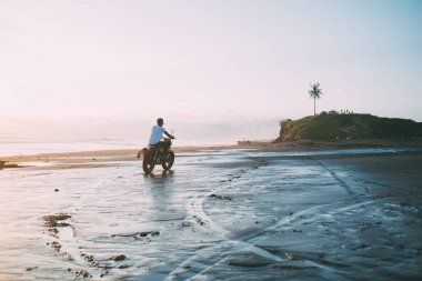 Yazın alacakaranlıkta deniz kenarında motosiklet süren beyaz tişörtlü erkek motorsikletlinin arka görüntüsü