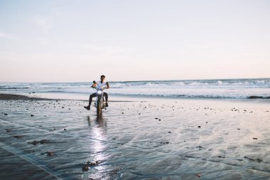 Gündelik giysiler ve güneş gözlüğü takan genç adam motosiklette oturmuş ıslak kumlu deniz kıyısında dinleniyor.