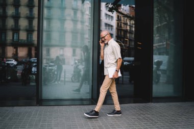 Şehirde elinde tabletle gezen ve cep telefonuyla konuşan, gündelik giyinmiş, gülümseyen yaşlı bir erkeğin yan görüntüsü