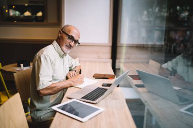 Resmi giyinen, gözlüklü, dizüstü bilgisayar ve tablet üzerinde çalışan ve modern kafede otururken not alan pozitif sakin girişimcinin yan görünüşü