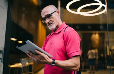 Şehirde boş zamanlarında kablosuz interneti taramak için dokunmatik ped kullanan yaşlı erkek blogcu, klasik göz giysisi giyen beyaz adam dijital tablette 4G 'ye bağlanan yayın haberleri okuyor