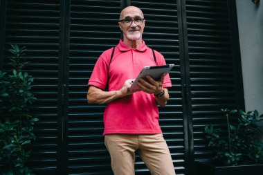 Modern dokunmatik bloknotu elinde olan, boş zamanlarında kameraya bakan, günlük pembe tişört giymiş, dijital tablette 4 g internet kullanan, olgun, beyaz bir adamın yarım boy portresi.
