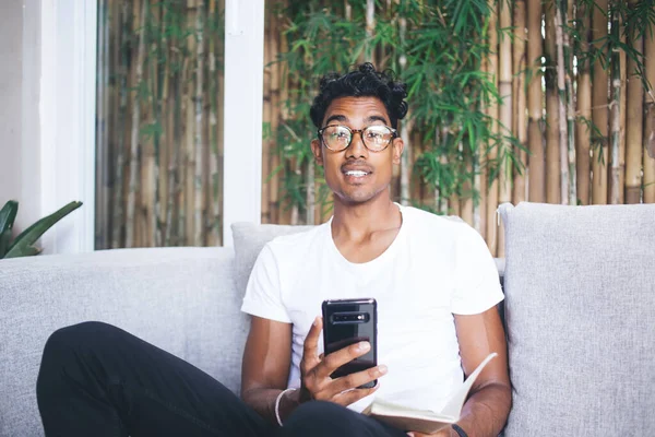 Zrelaksowany Czarny Włosy Młody Mężczyzna Okularach Notatnikiem Ręku Przeglądanie Smartfona — Zdjęcie stockowe