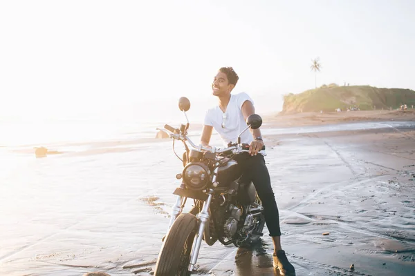 身穿休闲装 戴着太阳镜的成年自行车手坐在摩托车上 坐在湿沙滩上 在海景模糊的背景下拿着把手 一边看着相机 一边笑一边笑 — 图库照片
