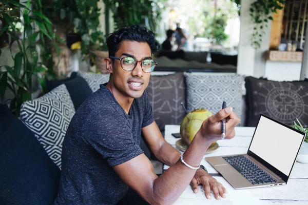Worried youthful Hispanic college student in glasses looking at camera while writing essay and sitting at table with empty screen laptop in opened cafe