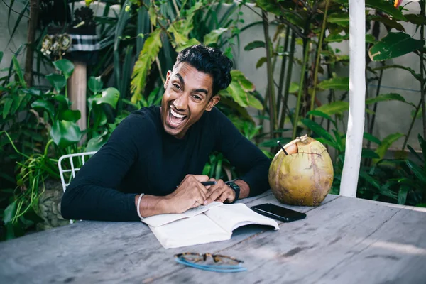 夏日快乐英俊的西班牙男人坐在餐桌旁 一边喝椰子 一边用手机 并在笔记本上写下自己的想法 — 图库照片