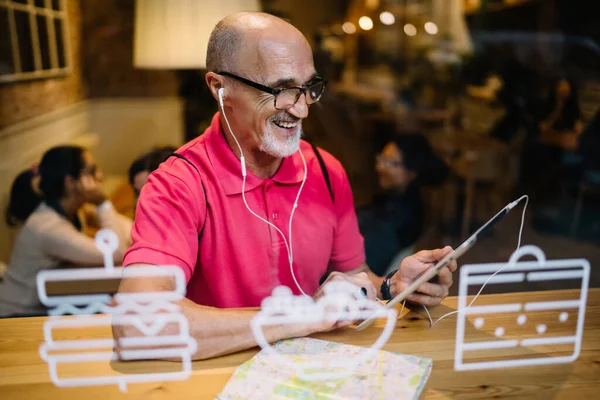 Cheerful senior in classic eyewear and electronic headphones using media application on modern touch pad for watching online video, happy man in earphones listening music while making web booking