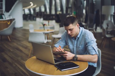 Konsantre olmuş erkek serbest çalışan modern bir kafede oturuyor ve bir yandan iş projesi üzerinde çalışırken bir yandan da not panosuna yazı yazıyor ve dizüstü bilgisayar kullanıyor.