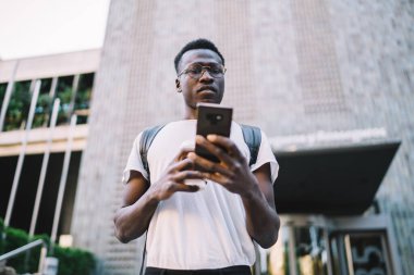 Düşük açılı afro-amerikan erkek beyaz gözlüklü, sırt çantalı ve cep telefonlu bina girişinde dikilirken