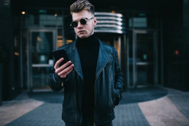 Siyah balıkçı yakalı ve ceketli genç adam ofis binasının önünde duruyor ve bilgi aramak için akıllı telefonu kullanıyor.