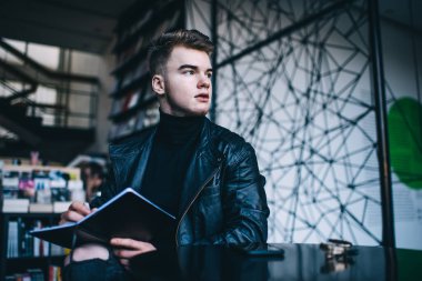 Siyah balıkçı yaka kazaklı ve deri ceketli, modaya uygun genç bir yazar kafeteryada oturmuş yeni bir makale yazarken başka tarafa bakıyor ve düşünüyor.