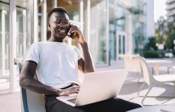 Smiling dark skinned hipster guy in eyewear for vision correction working remotely and talking on smartphone in roaming, positive african american male blogger on cafe terrace making mobile call