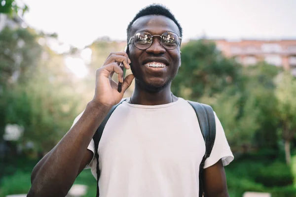 Cheerful black smiling guy with eyeglasses and backpack talking on phone and looking at camera against blurred green trees in city alley during sunny nice summer day