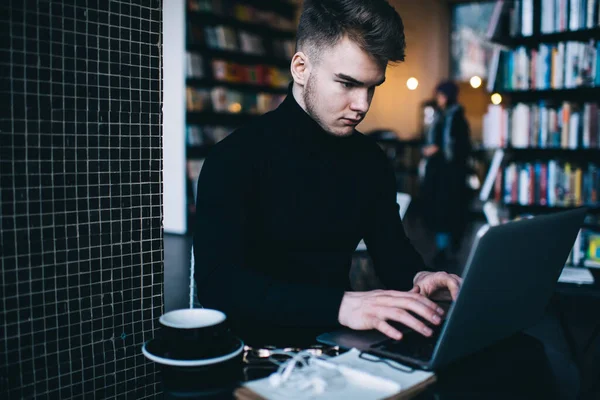Siyah balıkçı yakalı, kısa siyah saçlı, konsantre olmuş genç adam kütüphanede otururken bilgi edinmek için laptop kullanıyor.