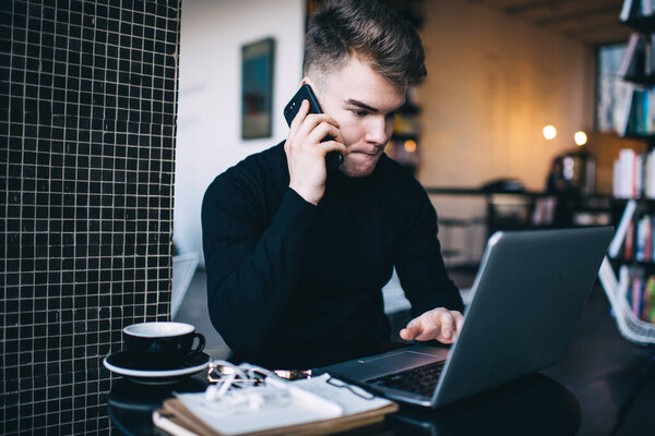 Focused young man in casual clothing having conversation on phone and pursing lips while working on laptop from cozy library