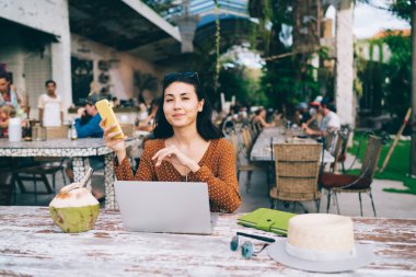 Bilgisayarlı, elinde cep telefonu olan Asyalı bir kadın. Kafede kameraya bakarken uzaktan iş projesi üzerinde çalışıyor.