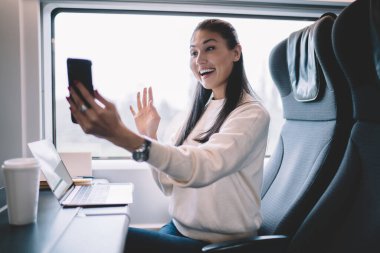Modern tren vagonunun penceresinin yanındaki rahat koltukta otururken gündelik kıyafetleriyle gülümseyen ve cep telefonuyla video görüşmesi yapan genç bir kadın.