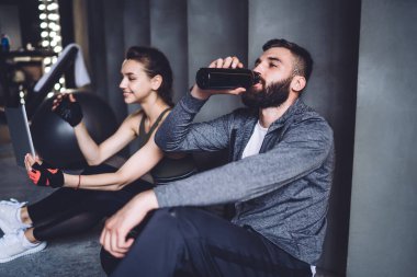 Beyaz kadın sporcu modern dijital tableti blog yazmak için kullanırken erkek arkadaşı antrenmandan sonra dinlenmek için su içiyor. Profesyonel kadın ve erkek antrenörler spor salonunda dinleniyor.