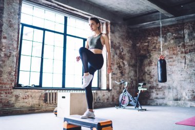 Tam vücut güçlü kadın fitness modeli siyah spor kıyafeti giyerek ve bacak kaldırarak spor salonundaki adım platformunda egzersiz yaparken.