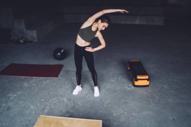 Spor elbiseli güçlü genç bir bayan ayakta ısınıyor spor salonunda gri renkte esneme egzersizi yapıyor.